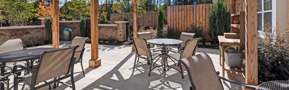 Outdoor patio area with multiple round tables and chairs under a wooden pergola, surrounded by a wooden fence and landscaped plants.