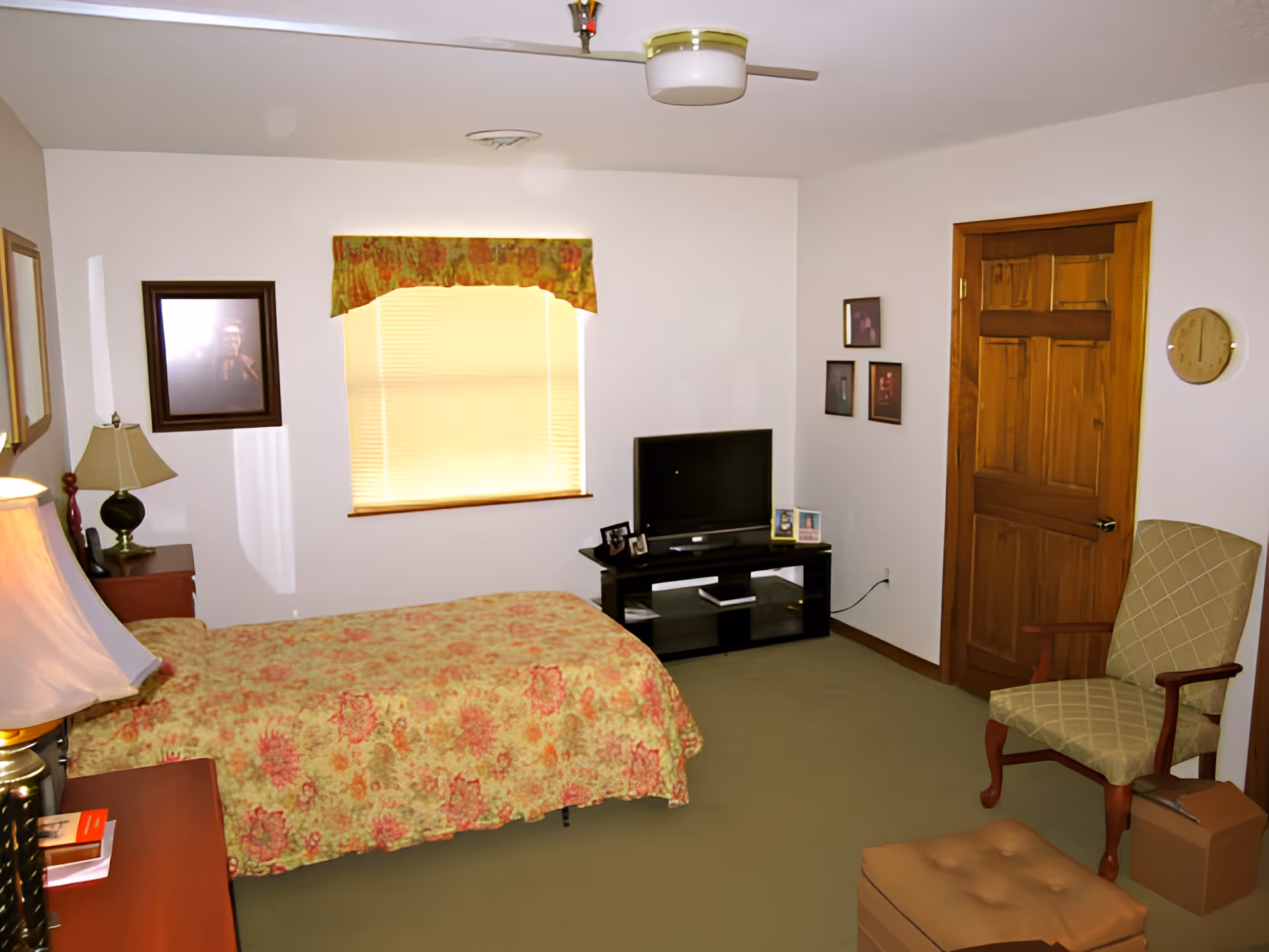 A bedroom with a single bed covered in a floral bedspread, a wooden door, a window with a valance, a TV on a black stand, a green upholstered chair, and a small ottoman. The walls are white and there are framed pictures hanging on the walls.