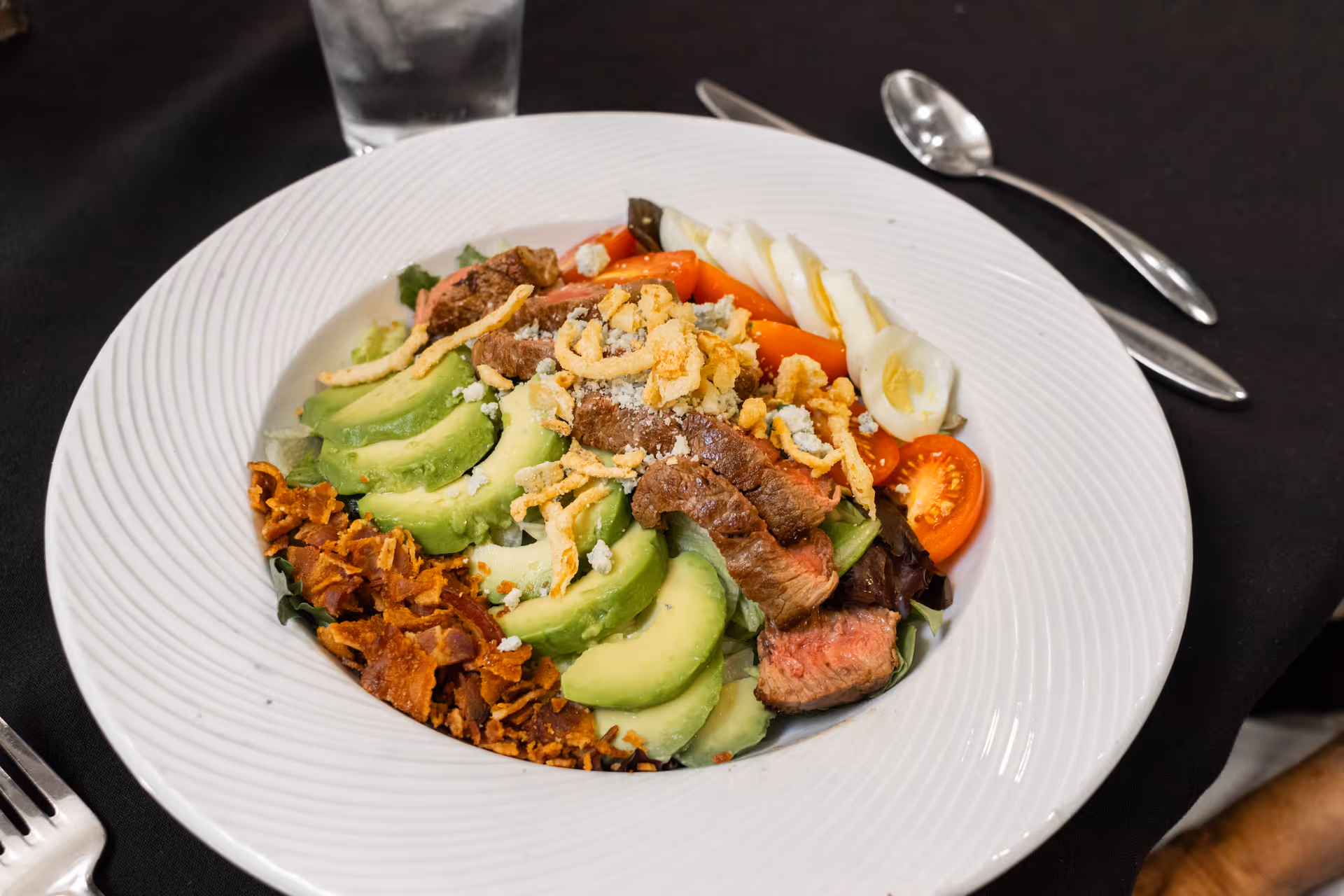 A white plate filled with a salad containing sliced avocado, cooked steak strips, hard-boiled egg slices, cherry tomatoes, bacon bits, and crispy fried onions on a bed of mixed greens. The plate is on a black tablecloth with a glass of water, a fork, and a spoon nearby.