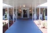 Covered entrance walkway leading to a glass door entrance of a building with stone walls and white pillars, flanked by two white rocking chairs and small landscaped areas with plants.