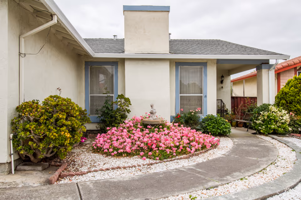Front exterior view of a single-story residential care home with beige walls and blue window frames. There is a curved concrete pathway leading to the entrance, surrounded by landscaped garden beds with pink flowers, green shrubs, and white decorative rocks. The sky is overcast.