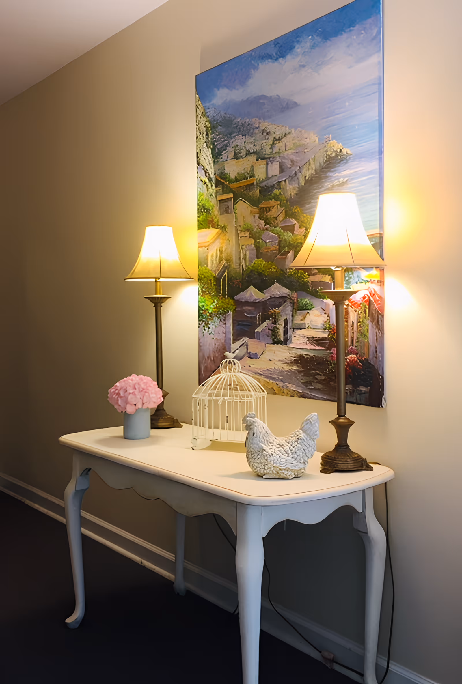 A white wooden console table against a beige wall with two lit table lamps, a small pink flower arrangement in a blue vase, a white decorative birdcage, and a white ceramic chicken figurine. Above the table hangs a large colorful painting depicting a coastal village scene.