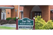 Exterior view of Forest Park Health Center & Rehabilitation building with a brick facade, arched entrance, and a green sign in front displaying the facility name. There are shrubs and greenery around the sign and building.