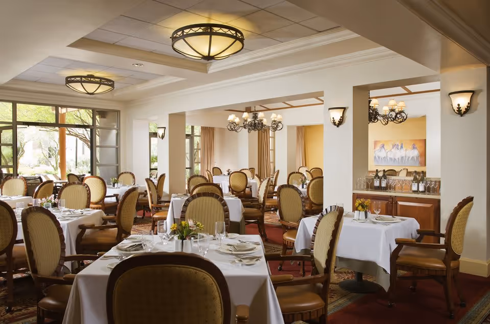 A spacious dining room with multiple tables covered in white tablecloths, each set with plates, glasses, and silverware. The room features elegant upholstered chairs, chandeliers, wall sconces, and large windows allowing natural light to brighten the space. There is a sideboard with wine bottles and glasses, and a painting of horses on the wall.