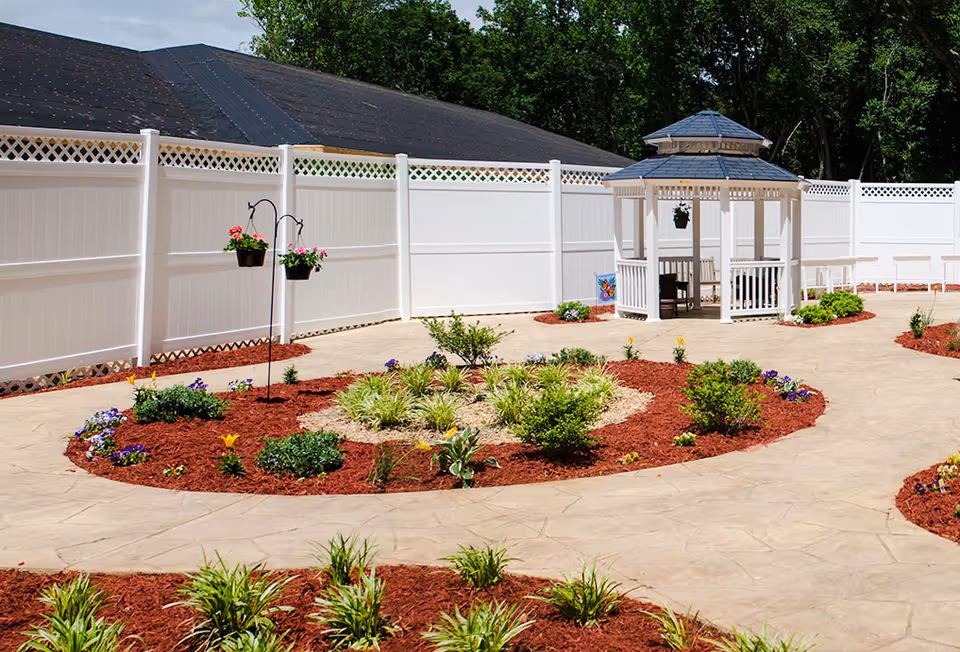 Outdoor garden area with a white gazebo, surrounded by a white fence and landscaped flower beds with various plants and flowers. There is a paved walkway curving through the garden.