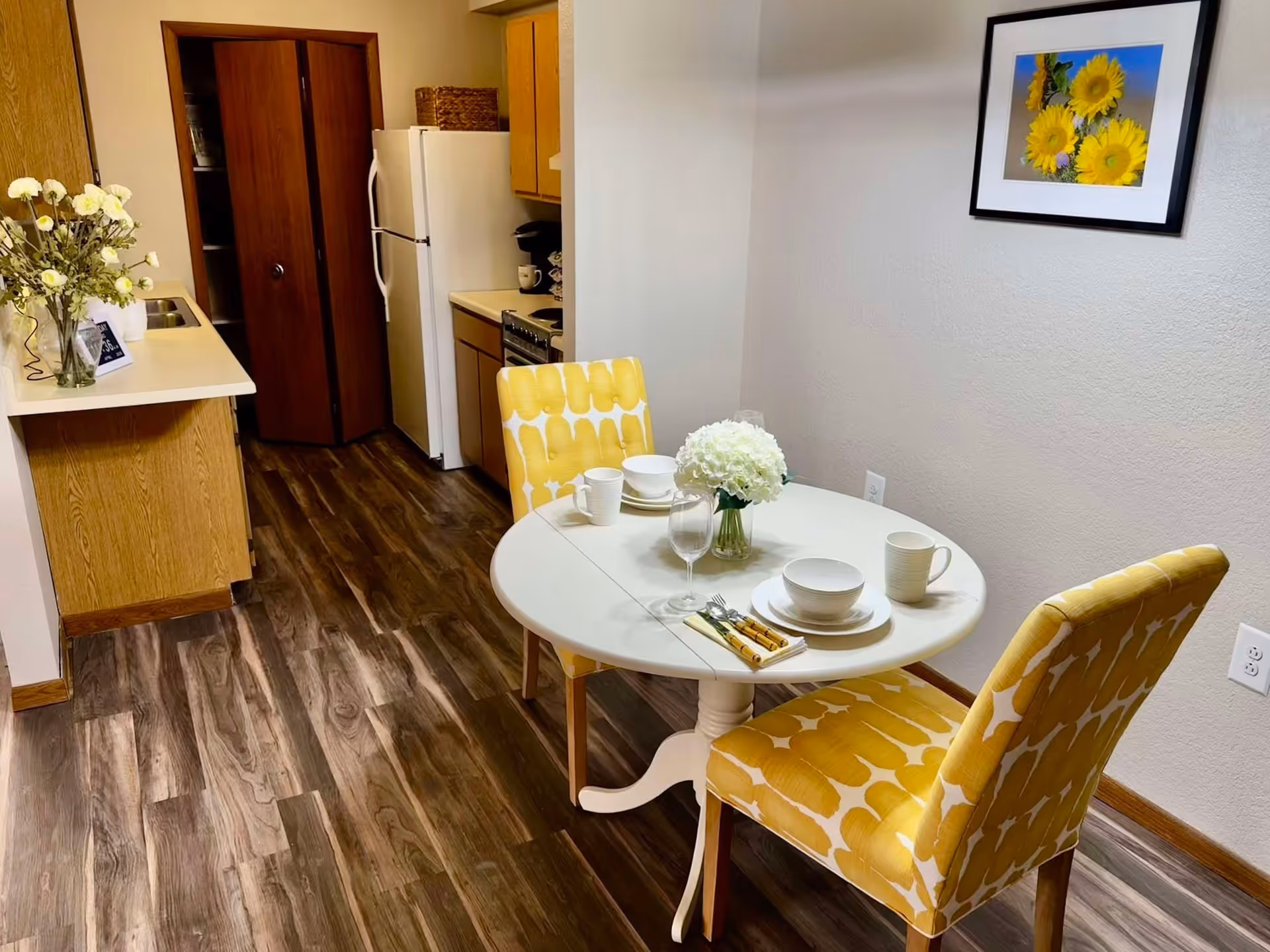 A small dining area with a round white table set for two, featuring white dishes, cups, and a vase with white flowers. Two yellow patterned chairs are positioned around the table. In the background, there is a kitchen with wooden cabinets, a white refrigerator, a stove, and a countertop with a vase of flowers. A framed picture of sunflowers hangs on the wall.