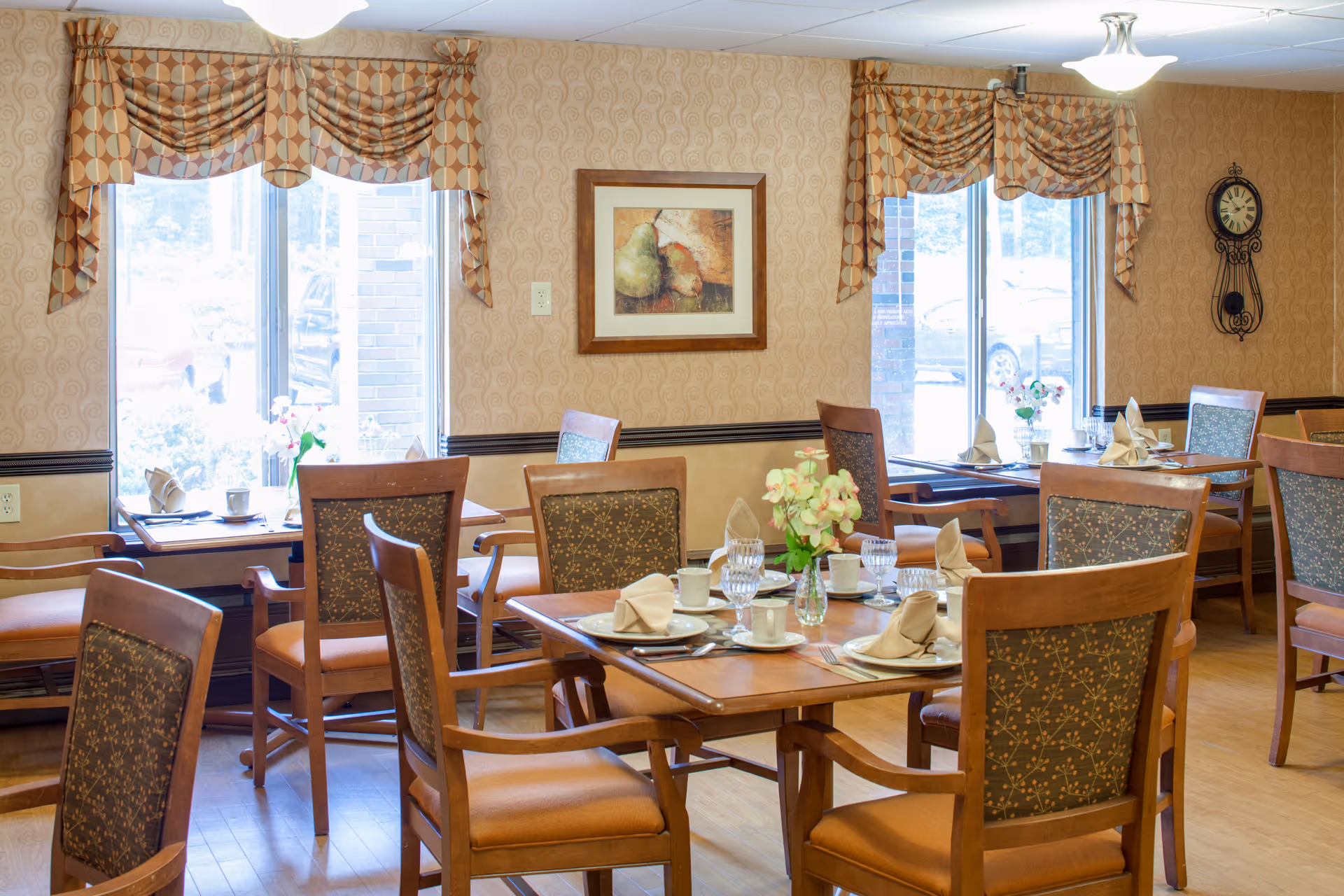 Bright dining room with multiple set wooden tables and chairs, floral centerpieces, and windows with patterned valances.