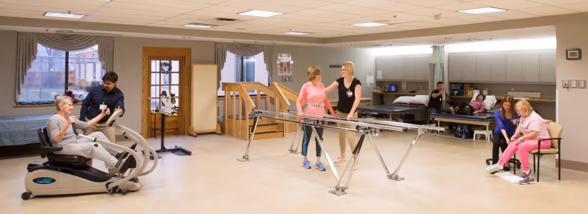 Several seniors and staff using exercise equipment and parallel bars in a rehabilitation therapy room.