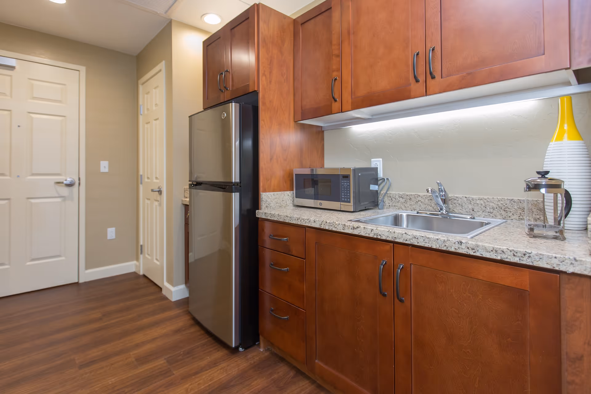 A kitchen area with wooden cabinets, a granite countertop, a stainless steel refrigerator, a microwave, a sink, and a decorative yellow and white vase next to a French press coffee maker. The floor is wooden and there are two closed doors in the background.