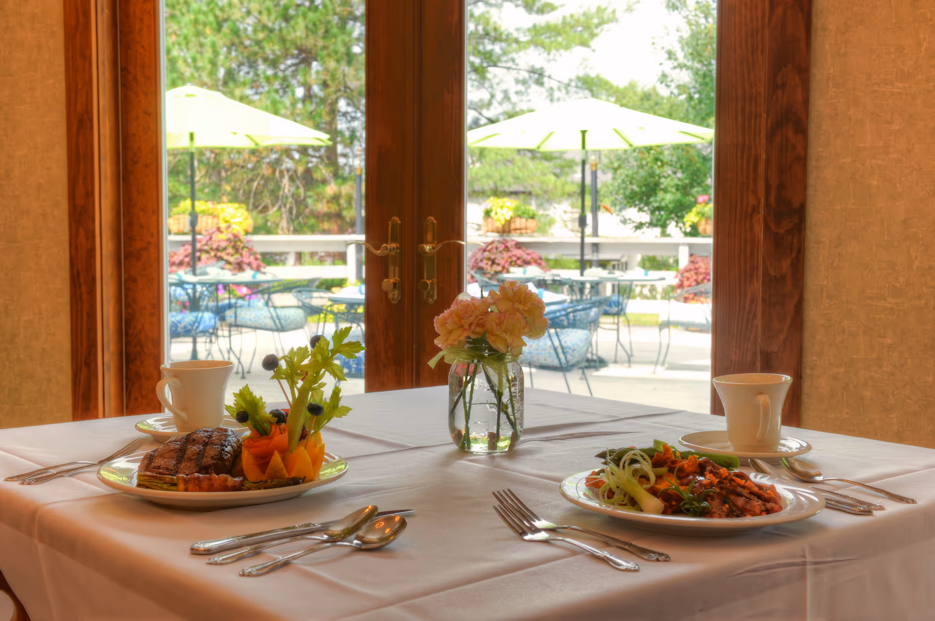 A dining table set for two with plates of food, cups, and silverware in front of large glass doors that open to an outdoor patio with tables, chairs, and green umbrellas.