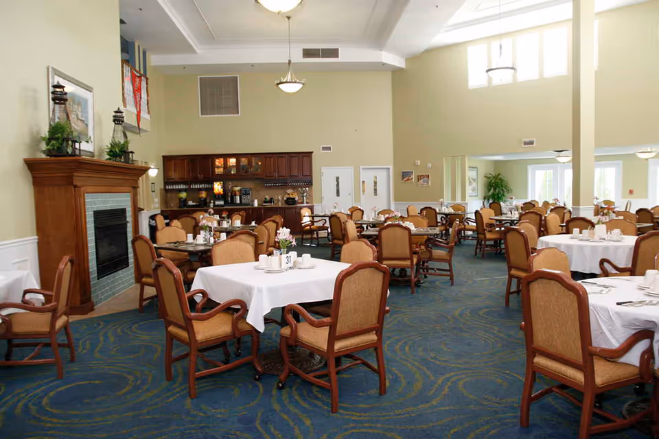 A spacious dining room with multiple tables covered in white tablecloths, each surrounded by cushioned wooden chairs. The room features a blue carpet with a swirling pattern, a wooden fireplace with decorative items on the mantel, and a cabinet with a coffee and beverage station. Large windows and high ceilings allow natural light to fill the space.