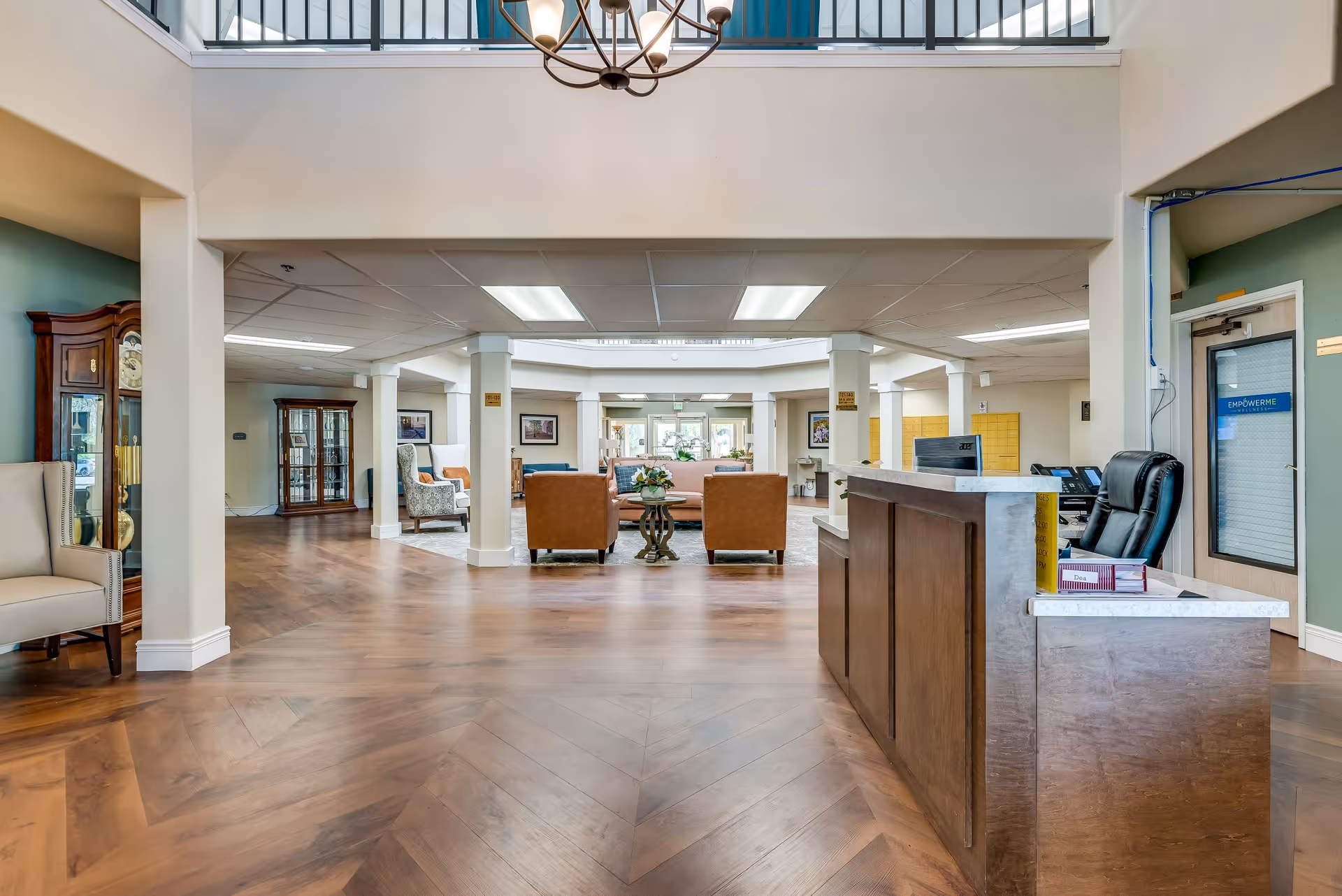 Spacious senior living facility lobby with wooden flooring, a reception desk on the right, comfortable seating area with leather chairs and a sofa in the center, and a grandfather clock on the left. The area is well-lit with ceiling lights and a chandelier, and there is a door labeled 'EmpowerMe Wellness' on the right side.