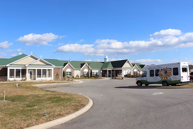 Exterior view of Meadow View Senior Living facility with a large paved driveway and a shuttle van parked near the entrance. The building has a green roof, beige walls, and multiple peaked roof sections under a blue sky with scattered clouds.