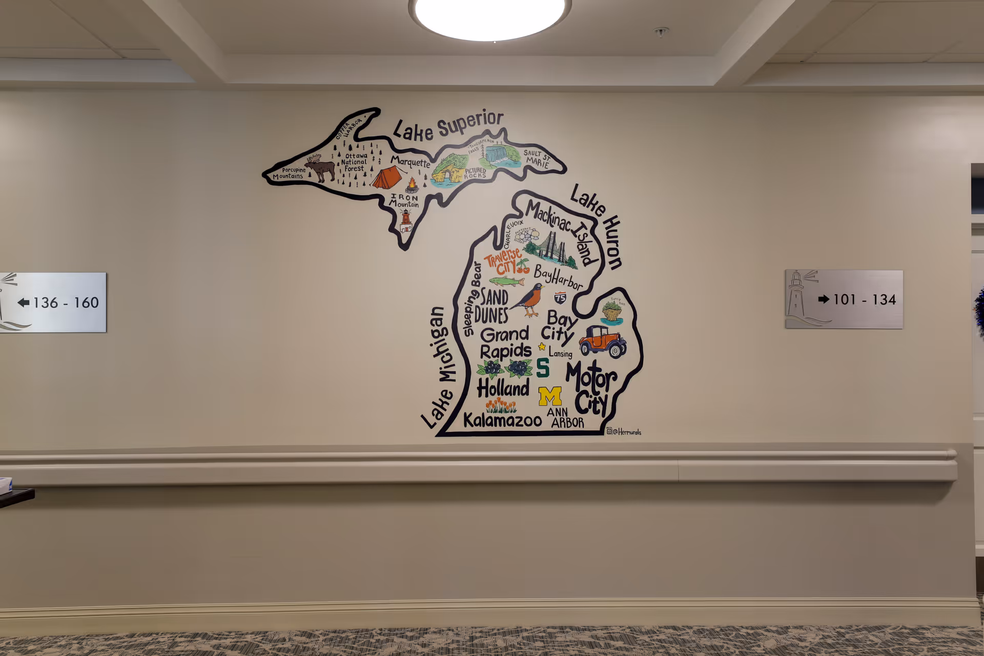 A hallway wall in a senior living facility featuring a colorful illustrated map of Michigan with notable locations and landmarks labeled. On either side of the map are directional signs indicating room numbers 136-160 to the left and 101-134 to the right. The wall is beige with a chair rail and carpeted floor.