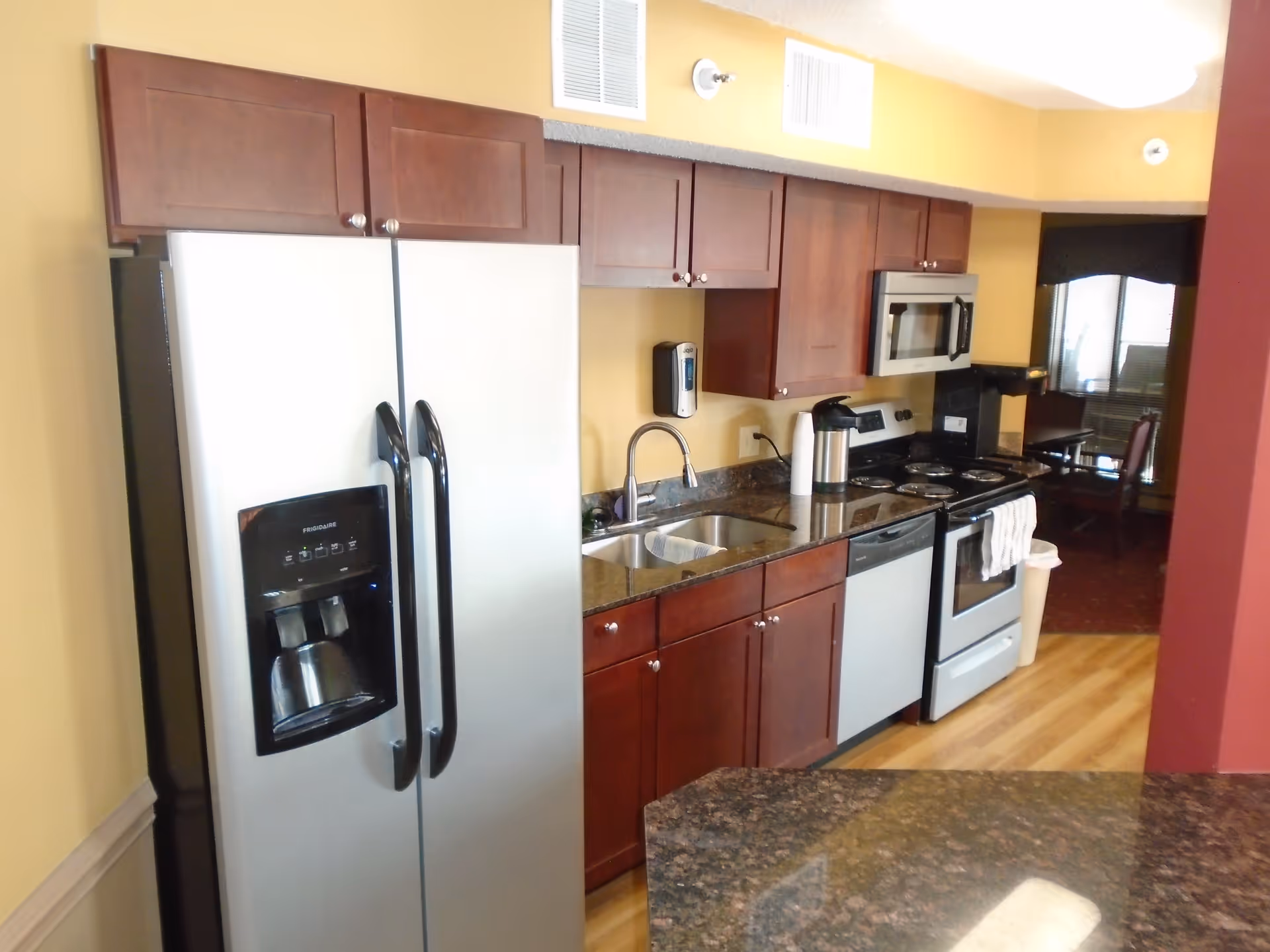 A kitchen area with wooden cabinets, a stainless steel refrigerator with water and ice dispenser, a sink with a faucet, a dishwasher, an electric stove with oven, a microwave above the stove, and a coffee maker. The kitchen has a granite countertop and wood flooring. In the background, there is a dining area with chairs and a window.