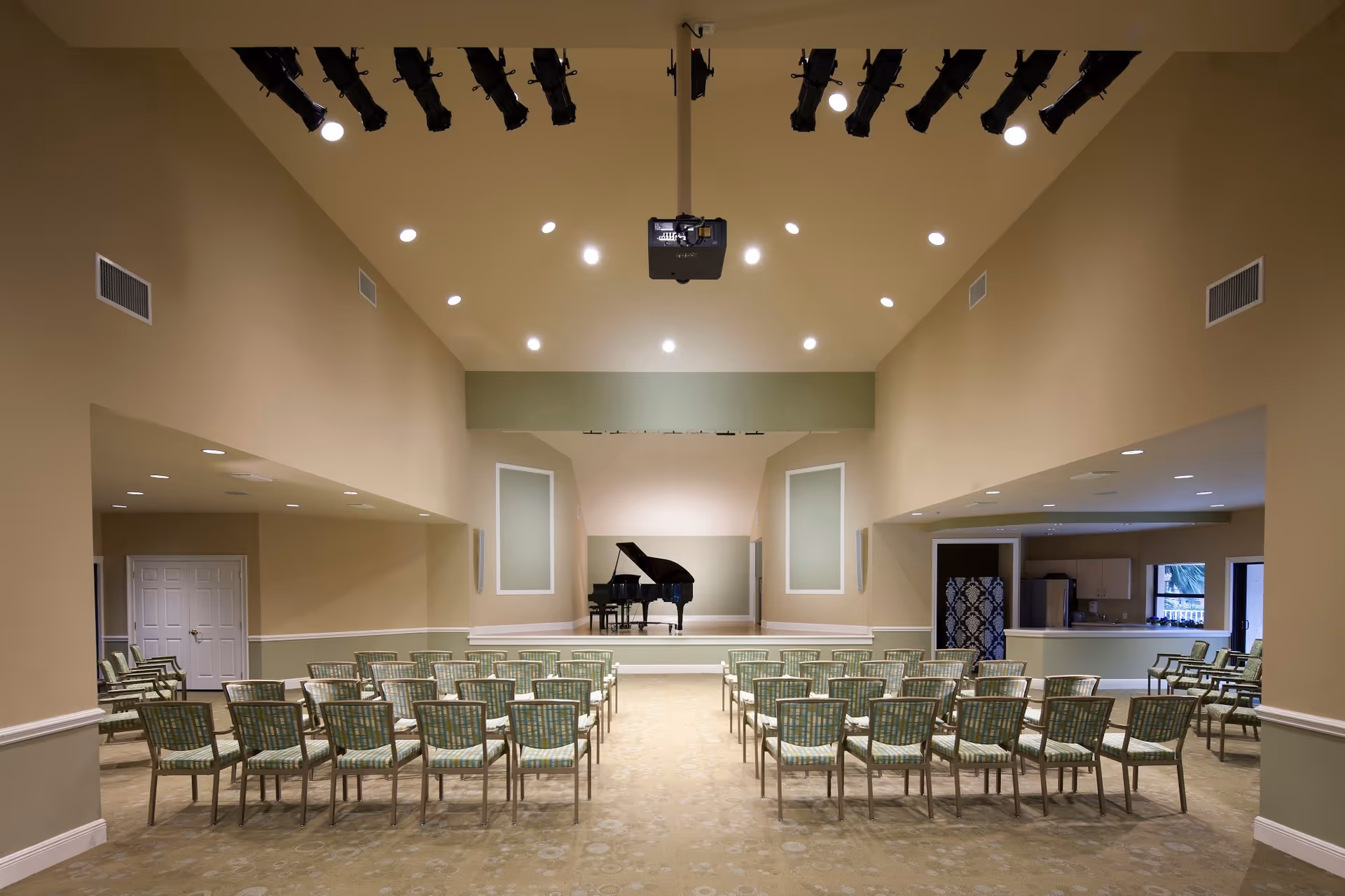 A spacious room with rows of green cushioned chairs facing a small stage with a grand piano. The room has beige walls, a high ceiling with multiple spotlights, and a projector mounted in the center. There are doorways and windows visible on the sides.