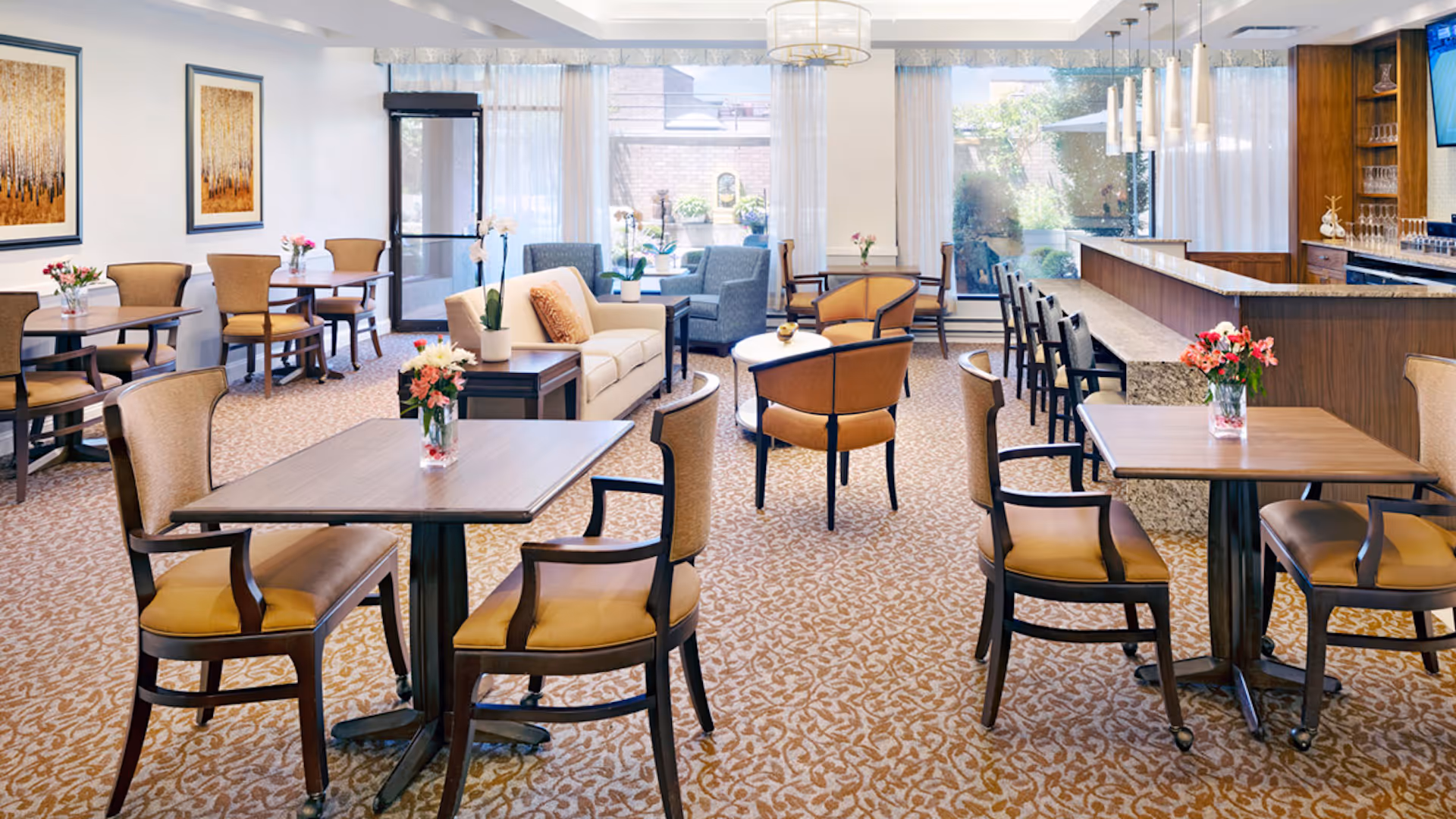 A bright and spacious dining and lounge area with multiple wooden tables and chairs, a beige sofa, two blue armchairs, and a bar counter with high stools. The room has large windows with sheer curtains allowing natural light to fill the space, and there are floral arrangements on the tables. Two framed artworks hang on the wall, and a television is mounted on the right side above the bar.