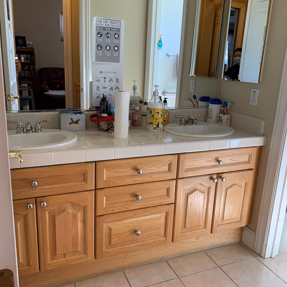 Double-sink bathroom vanity with oak cabinets, tiled countertop, toiletries, a paper towel roll, and a mirror reflecting a person and a COVID-19 hygiene poster.