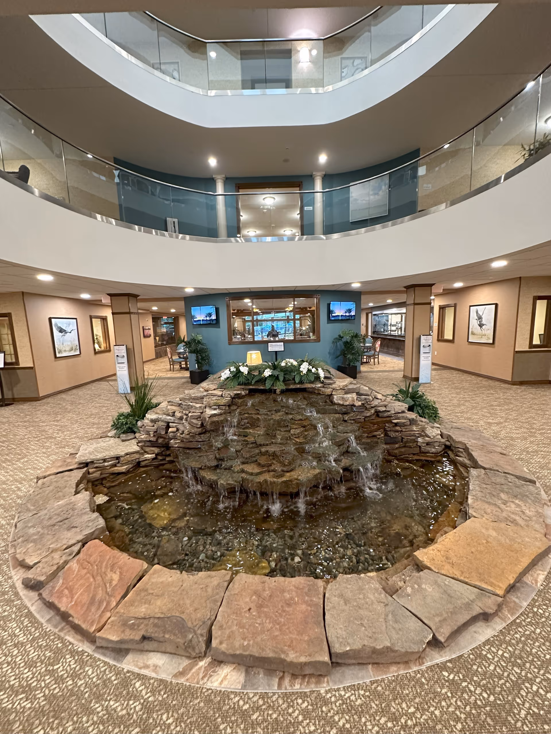Interior view of a spacious retirement community lobby featuring a central stone water fountain with flowing water and decorative plants. The lobby has a carpeted floor, beige walls adorned with framed artwork, and a two-level open ceiling with glass railings. There are seating areas and two wall-mounted televisions visible in the background.