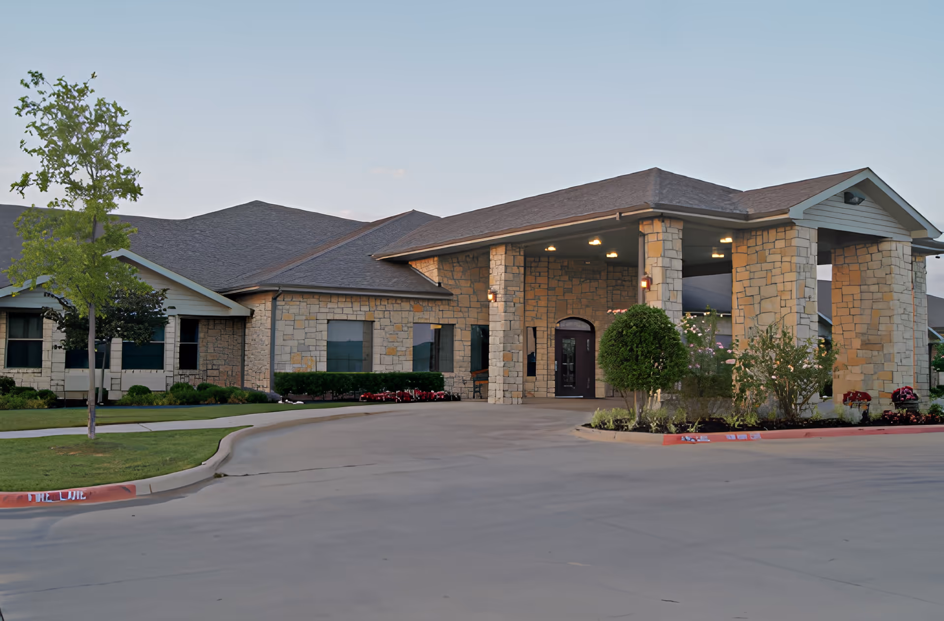 Stone-faced single-story senior living facility with a covered porte-cochere entrance, landscaped shrubbery, and a curved driveway.
