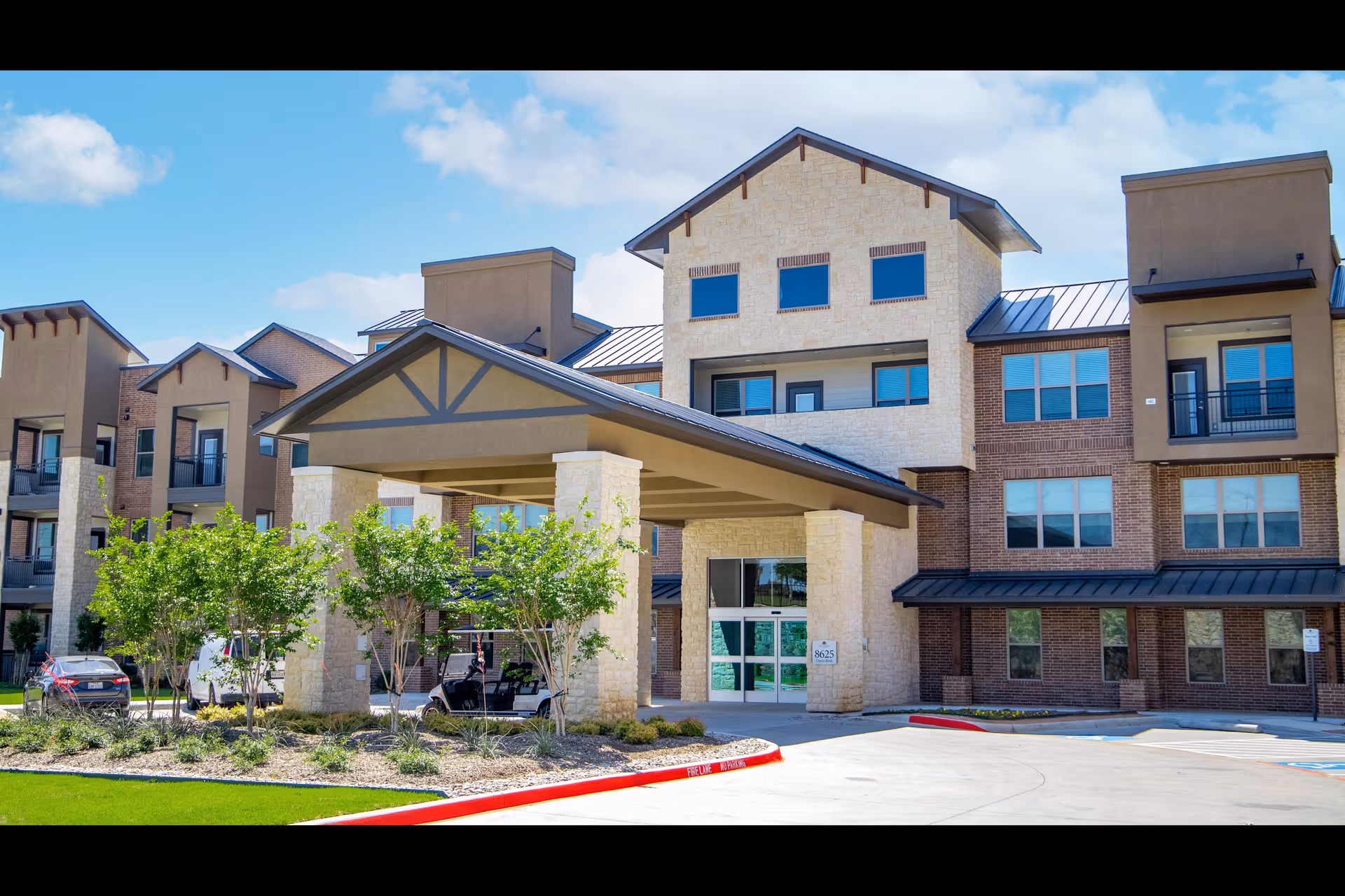 Exterior view of a modern senior living facility building with a covered entrance supported by stone pillars, surrounded by small trees and landscaping. The building has multiple floors with large windows and a mix of brick and stone facade under a blue sky with some clouds.