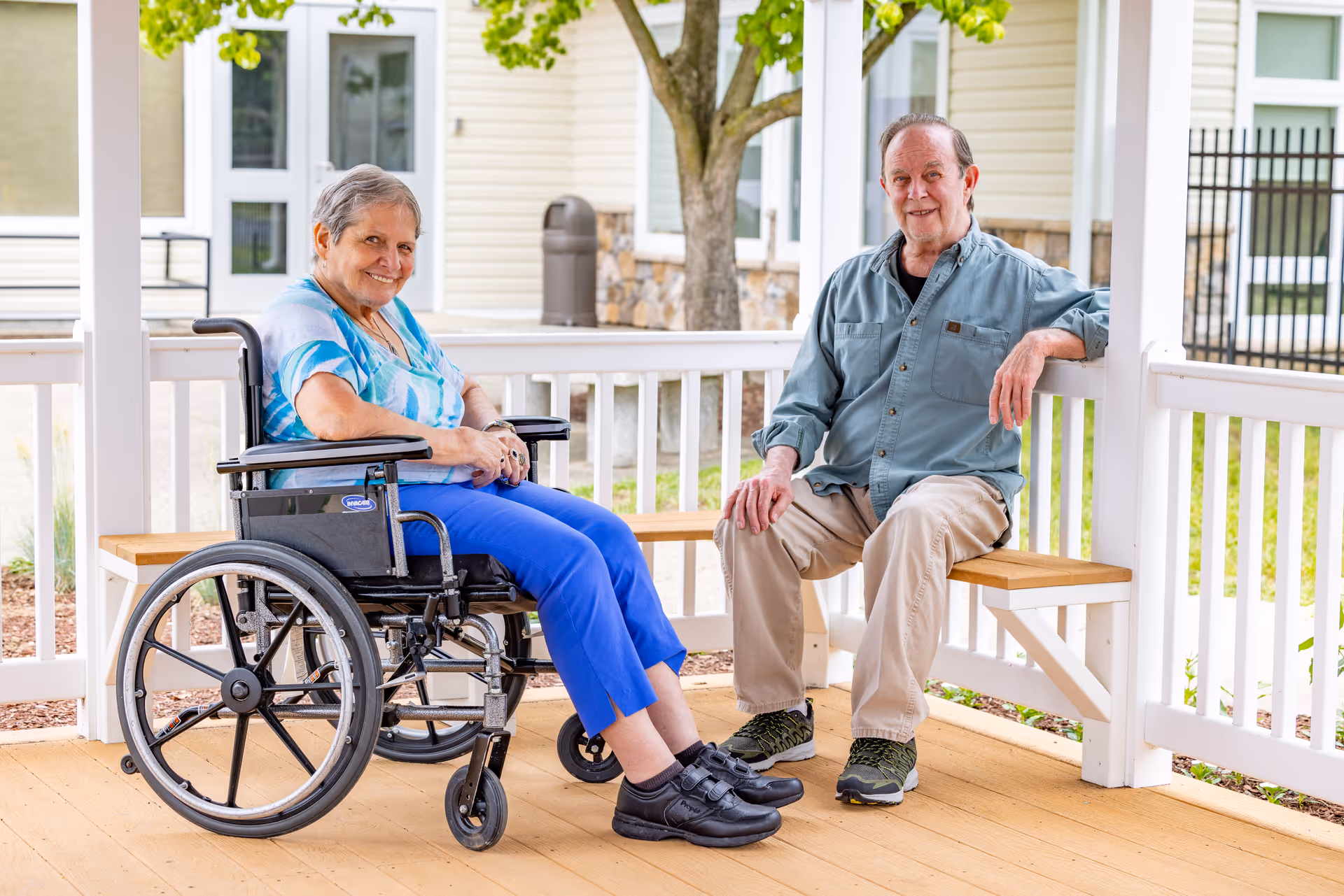 An elderly woman in a wheelchair and an elderly man sitting on a wooden bench under a covered outdoor patio area. They are smiling and appear to be enjoying the outdoor space of a senior living facility with a tree and building visible in the background.