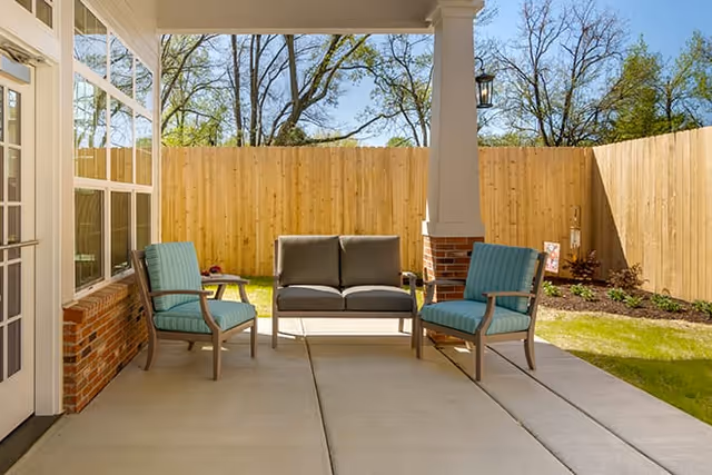 Covered outdoor patio area with a concrete floor, featuring a cushioned loveseat and two cushioned armchairs arranged around a small table. The patio is enclosed by a wooden fence, with trees visible in the background under a clear blue sky.