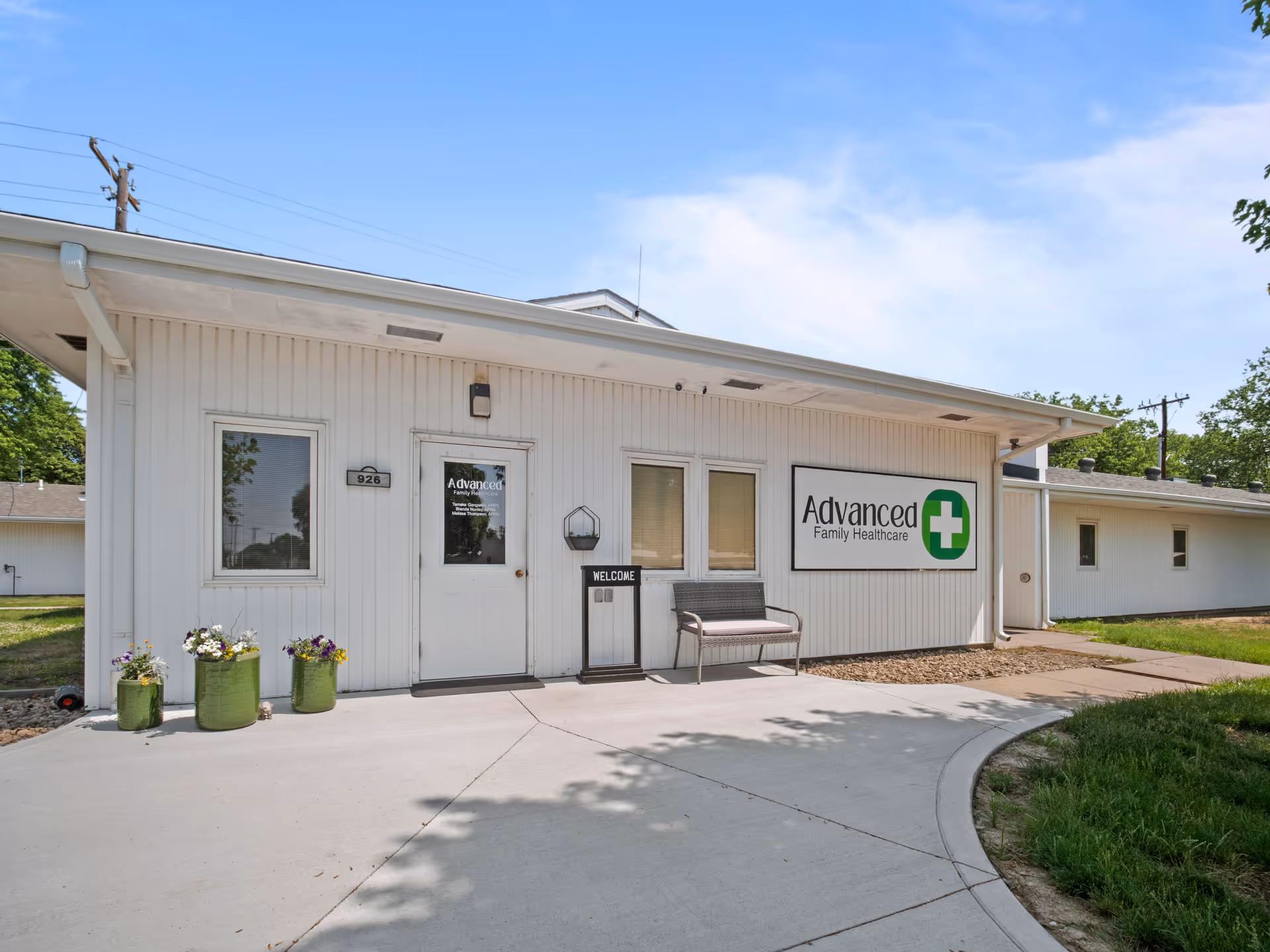Exterior view of a single-story white building with a sign that reads 'Advanced Family Healthcare'. There is a door with the number 926, two windows, a bench, a welcome sign, and three green planters with flowers near the entrance. The building is surrounded by a concrete walkway and some grass, under a clear blue sky.