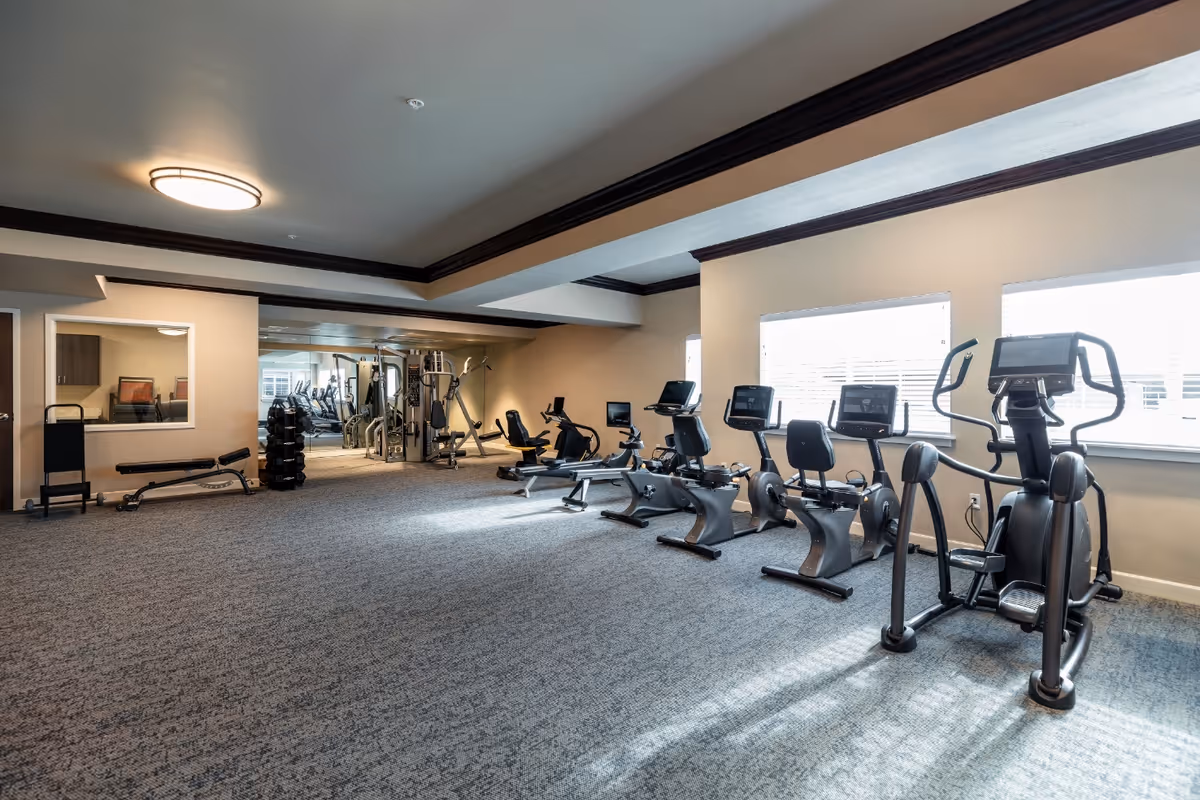 A spacious fitness room with exercise equipment including stationary bikes, an elliptical machine, weight machines, and free weights. The room has large windows letting in natural light, carpeted floors, and a ceiling light fixture.