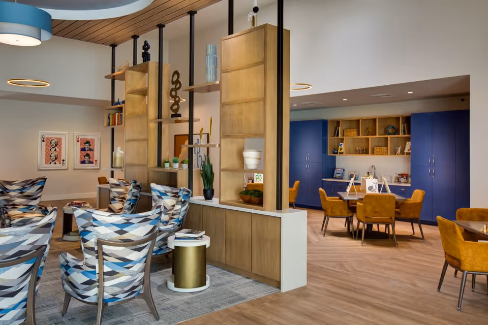 A modern senior living common area featuring patterned armchairs arranged around small tables on a rug. A wooden and metal shelving divider separates the seating area from a space with blue cabinets, open shelves, and a table with mustard yellow chairs. The room has warm wood flooring, contemporary lighting fixtures, and decorative artwork on the walls.