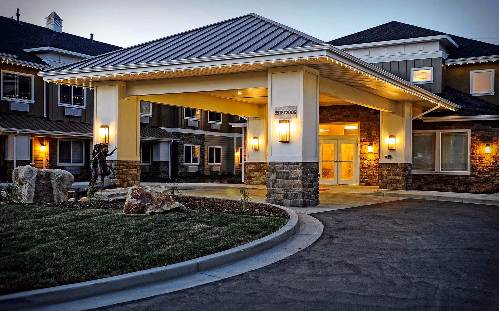 Covered main entrance of a senior living facility at dusk with lit lanterns, stone columns, landscaping and a small statue.