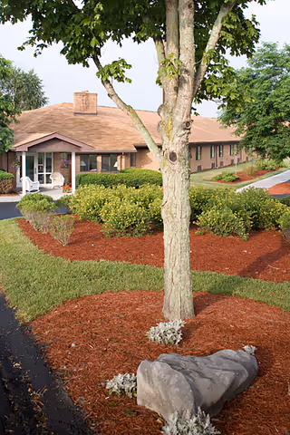 Front exterior of a single-story brick senior living building with a landscaped entrance, tree, and mulch beds.