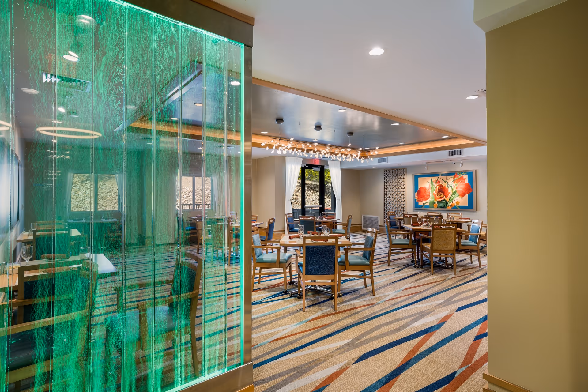 A spacious dining room with multiple wooden tables and chairs arranged neatly. The room features a colorful patterned carpet, a modern chandelier, large windows with white curtains, and a vibrant floral painting on the wall. A green illuminated glass panel is visible on the left side of the image.