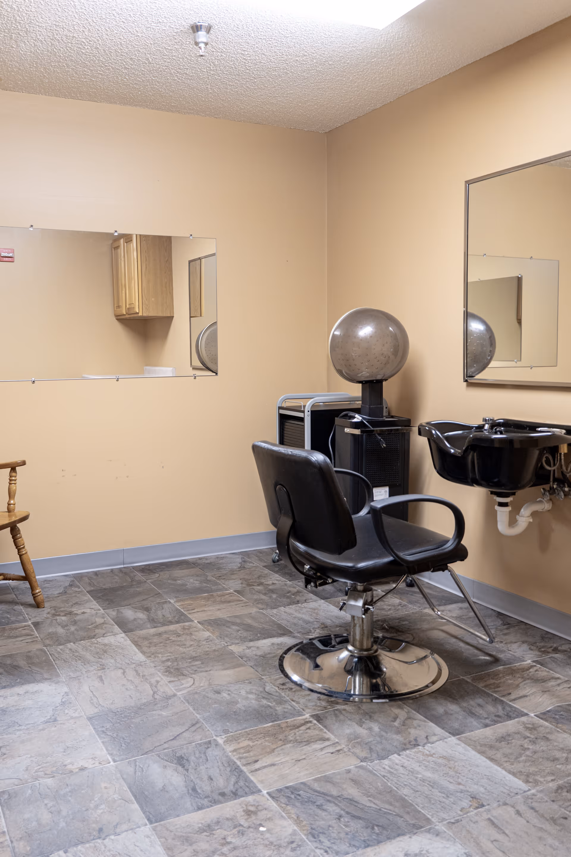 Interior of a salon room with a black salon chair, a hair dryer hood, a black wash basin attached to the wall, two large mirrors, a wooden chair, and beige walls with tiled flooring.