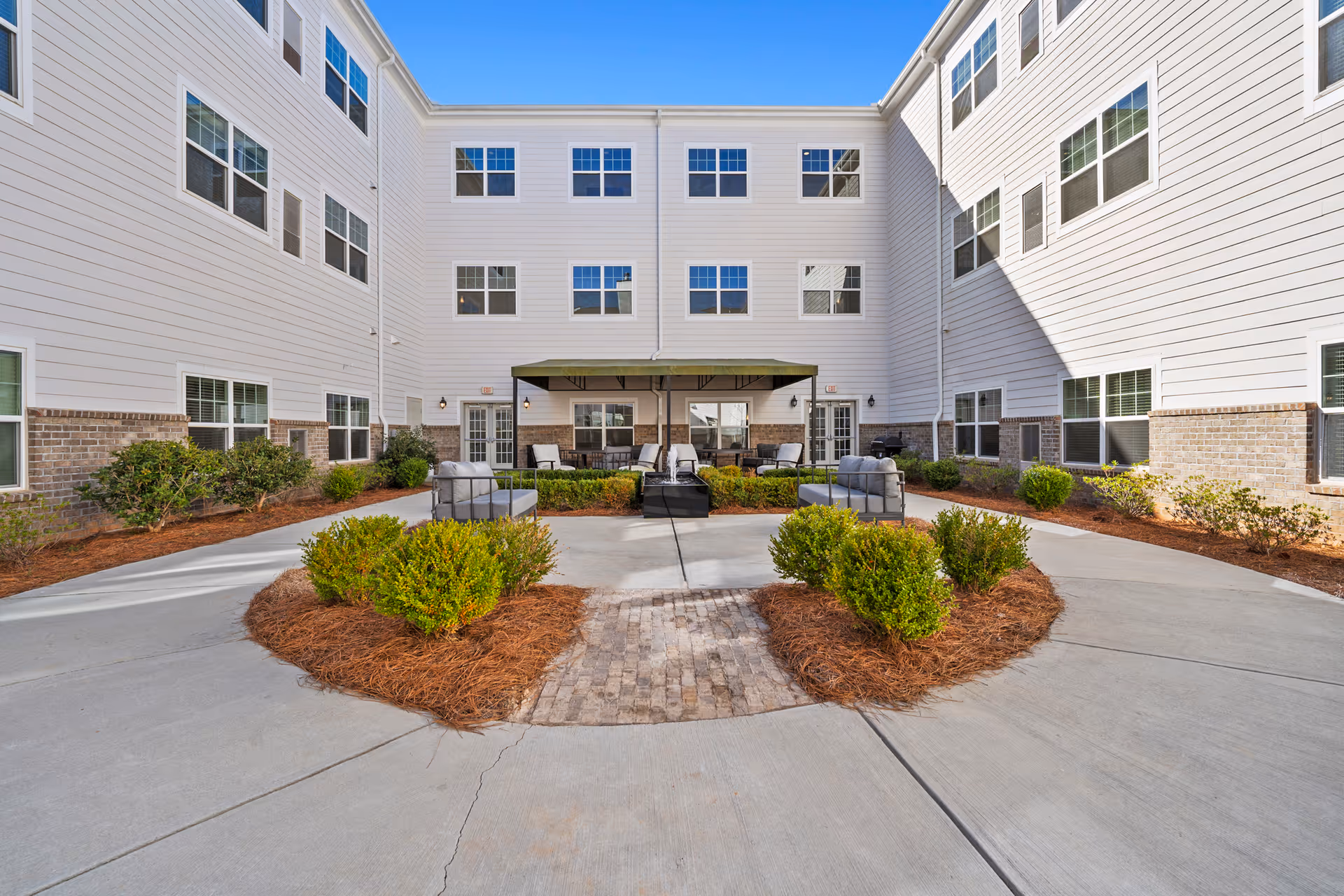Outdoor courtyard area of a senior living facility with white siding and brick exterior walls. The courtyard features a paved walkway, landscaped bushes, and outdoor seating with cushioned chairs arranged around a fire pit under a canopy.