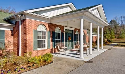 Front entrance of a single-story red brick building with a covered columned porch, chairs, and landscaped flowerbeds.