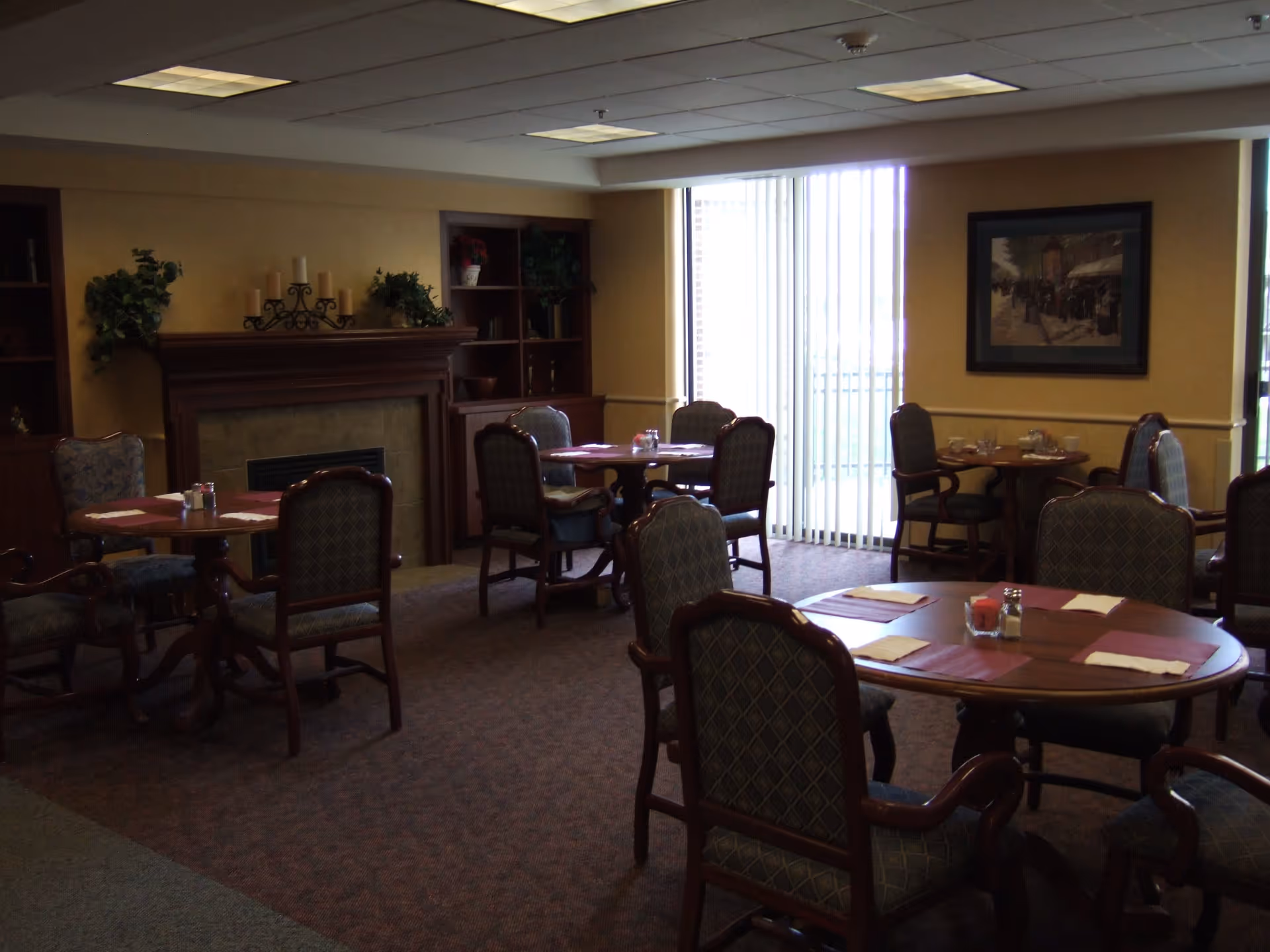 A dining room in an assisted living facility with several round wooden tables and cushioned chairs arranged around them. Each table has placemats, napkins, and condiments. There is a fireplace with candles and plants on the mantle, built-in shelves with decorative items, and a large window with vertical blinds letting in natural light. A framed picture hangs on the wall.