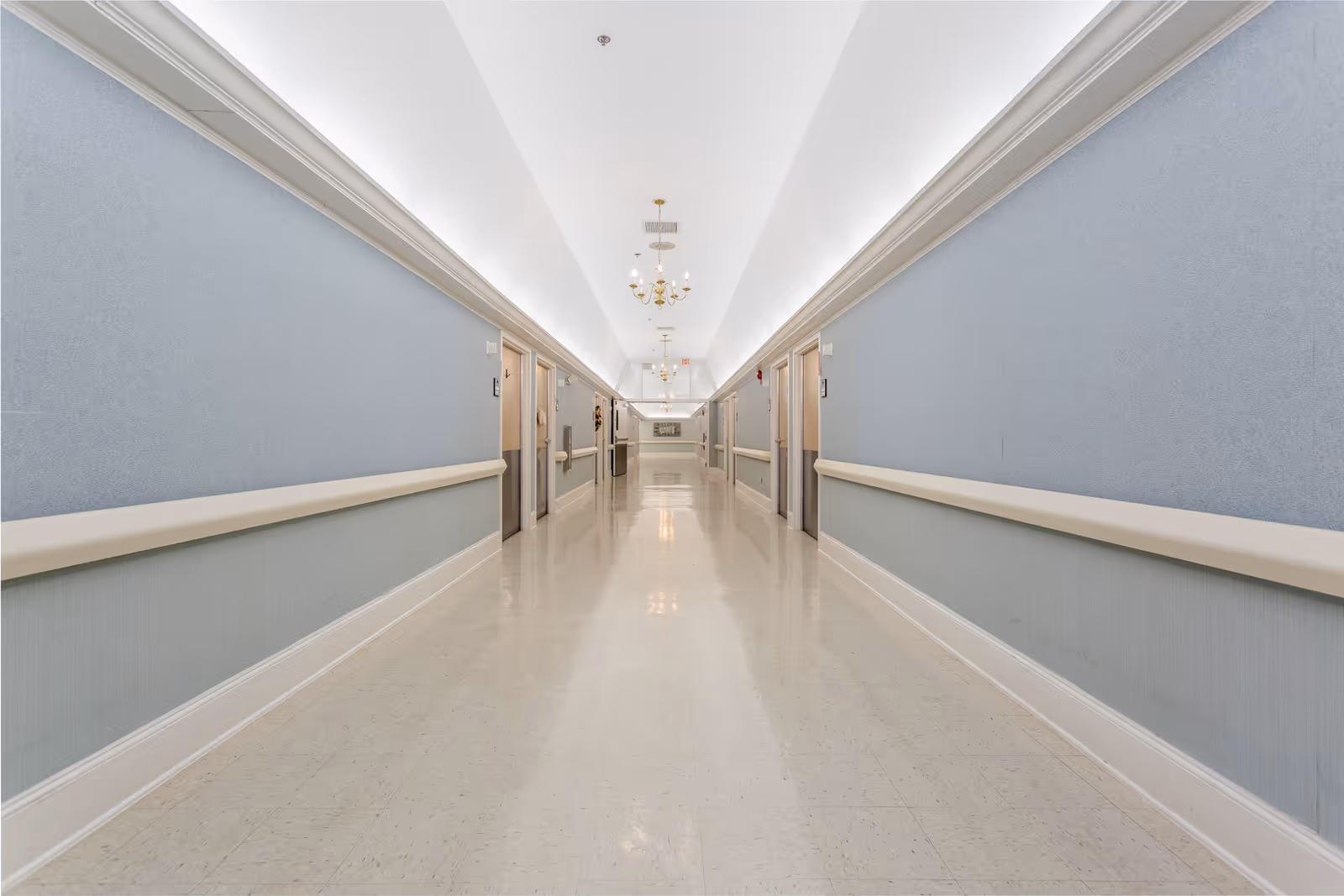 A long, clean hallway in a nursing and rehab facility with light blue walls, beige handrails, and multiple closed doors on both sides. The ceiling is white with chandeliers and recessed lighting, and the floor is a polished light-colored tile.