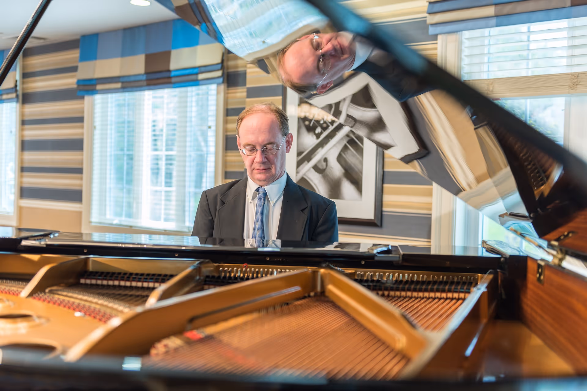 A man in a suit and tie is playing a grand piano in a room with striped wallpaper and large windows with blinds. The piano's open lid reflects the man's face. There is a black and white framed photo on the wall behind him.