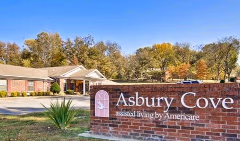 Exterior view of Asbury Cove assisted living facility with a brick sign in the foreground displaying the facility name and a single-story building with a driveway and trees in the background.