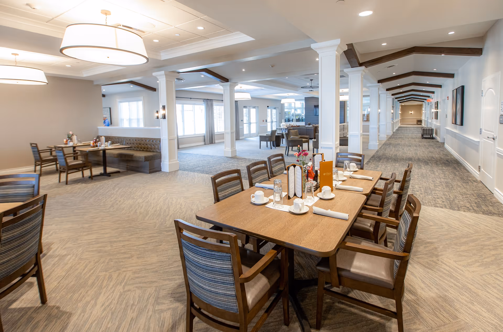 A bright and spacious dining area in a senior living facility with wooden tables and chairs. One table is set with cups, glasses, napkins, menus, and condiments. The room features large windows with curtains, white columns, and modern ceiling lights. A long hallway extends to the right with artwork on the walls.
