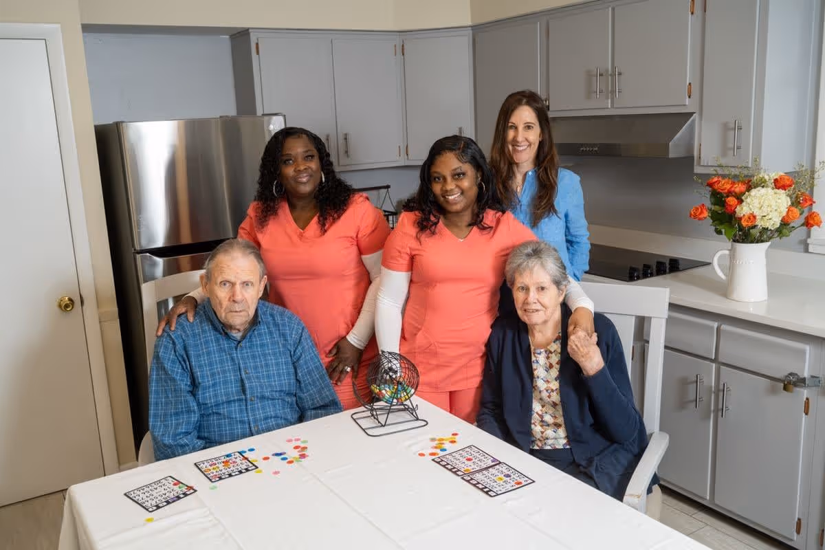 Two elderly individuals seated at a table with bingo cards and markers, accompanied by three smiling caregivers standing behind them in a kitchen setting with white cabinets, a stainless steel refrigerator, and a vase of flowers on the counter.