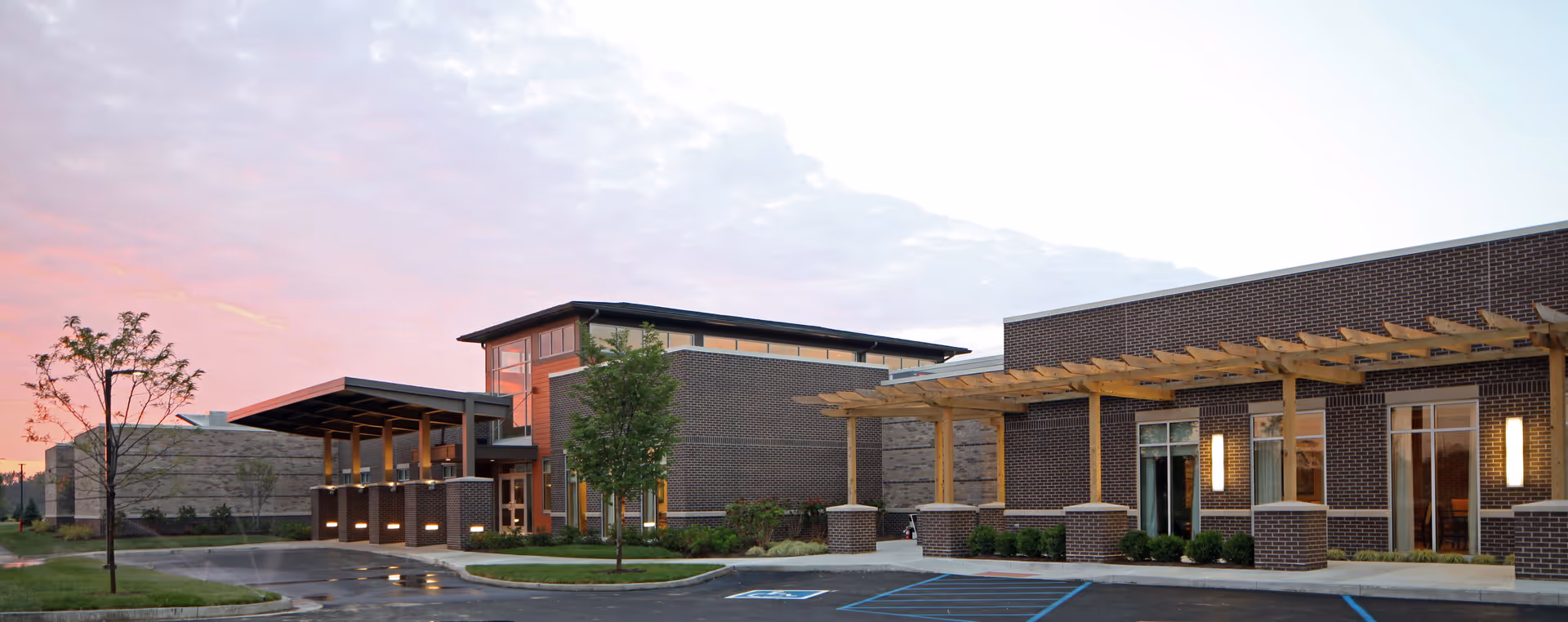 Exterior view of a modern brick building with large windows and a wooden pergola over the entrance area, surrounded by small trees and landscaping, under a partly cloudy sky at sunset.