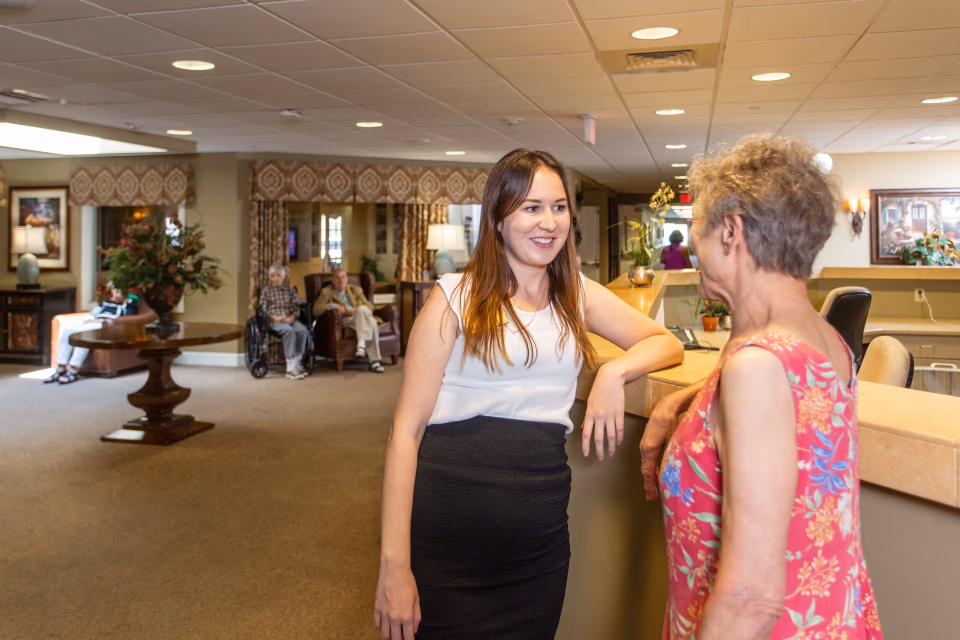 Two women engaged in conversation at a reception desk inside a senior living facility. In the background, elderly residents are seated in a common area with armchairs and a round wooden table with a floral arrangement. The room is warmly lit with lamps and decorated with framed artwork and patterned curtains.