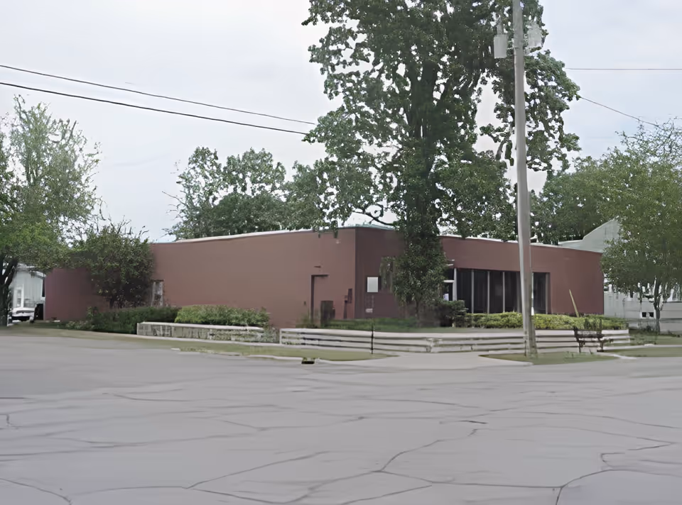 Single-story red-brick institutional building with large trees, a low stone planter and a utility pole in front.