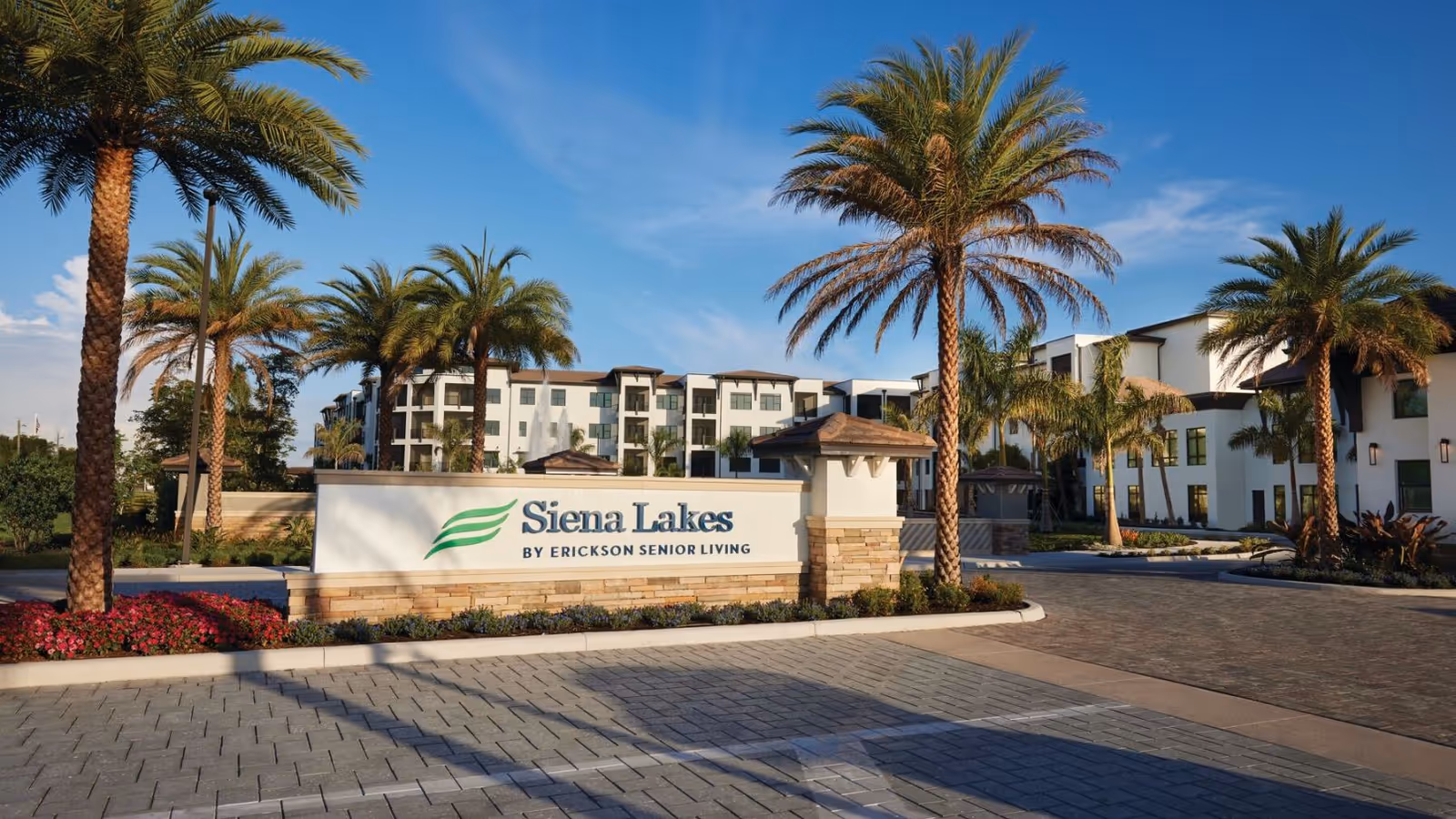 Entrance sign reading 'Siena Lakes' in front of palm trees and the senior living building under a blue sky.