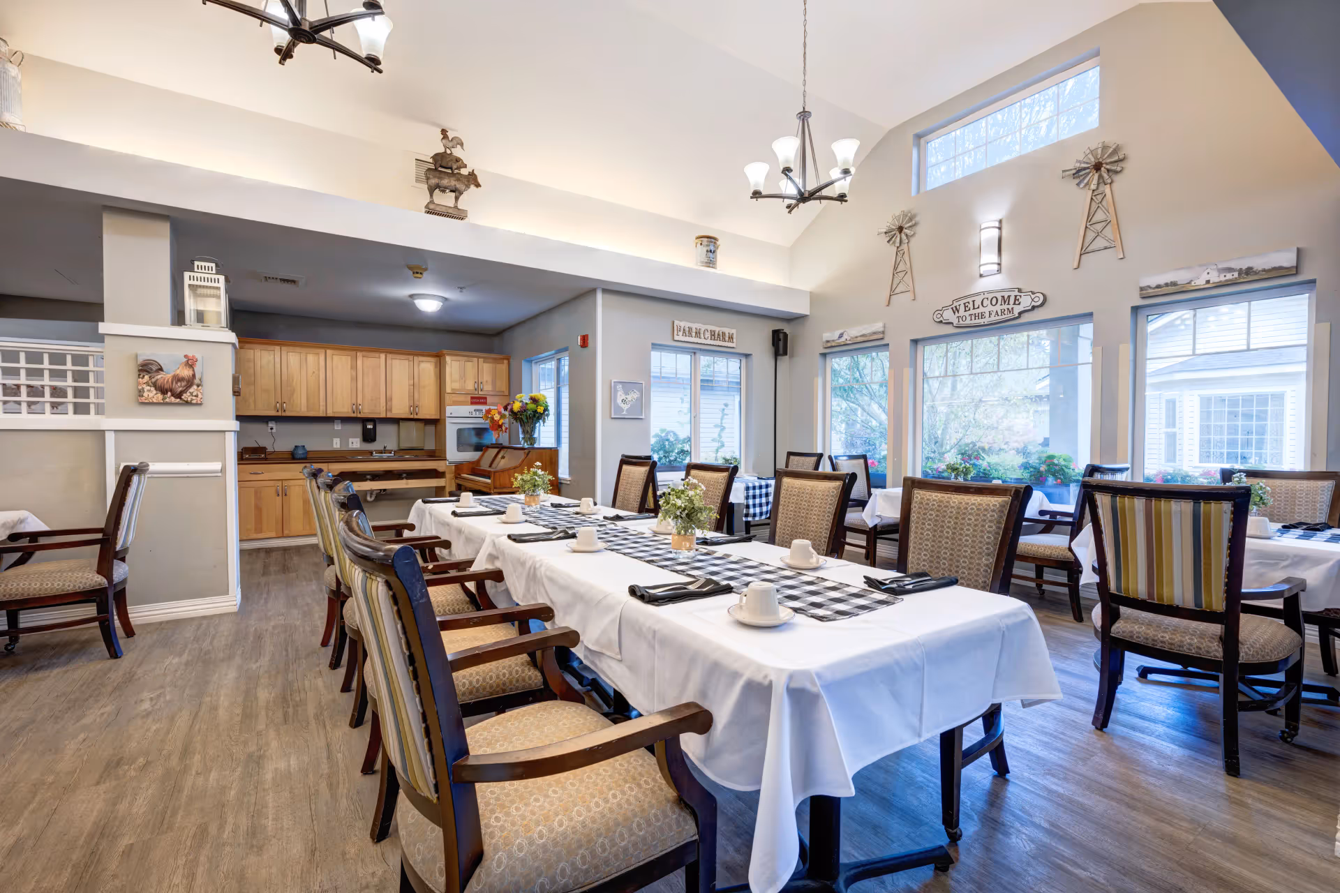 Bright dining room with a long table set for a meal, multiple chairs, and a kitchenette in the background.
