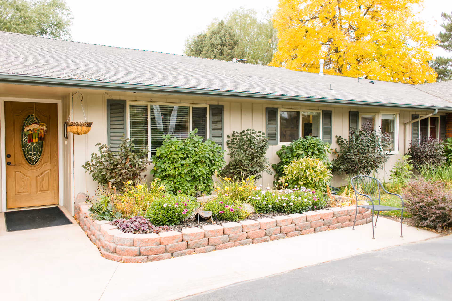 Front of a single-story assisted living building showing a wooden entry door, windows with shutters, a raised flowerbed, and a metal chair on the walkway.