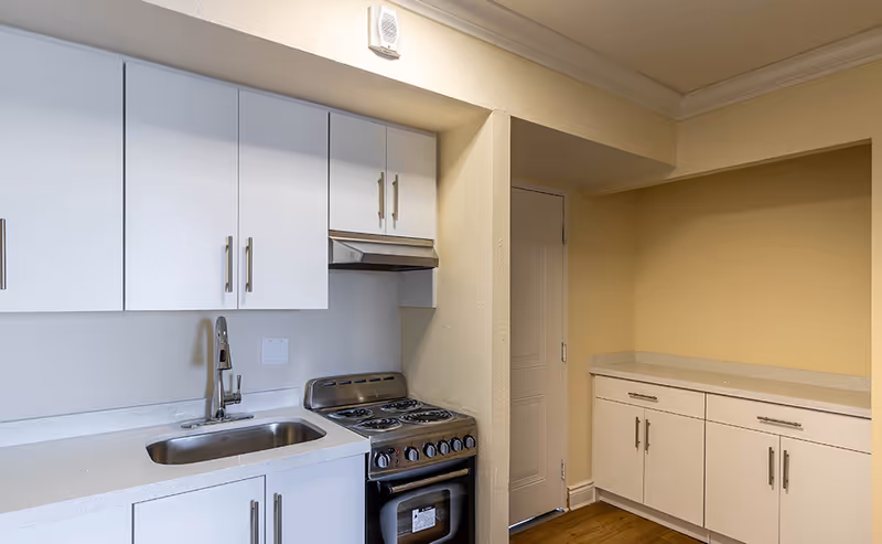A clean kitchen area with white cabinets, a stainless steel sink, an electric stove with four burners, and a countertop. There is a door next to the stove and additional cabinets with a countertop on the right side. The walls are painted beige and the floor is wooden.
