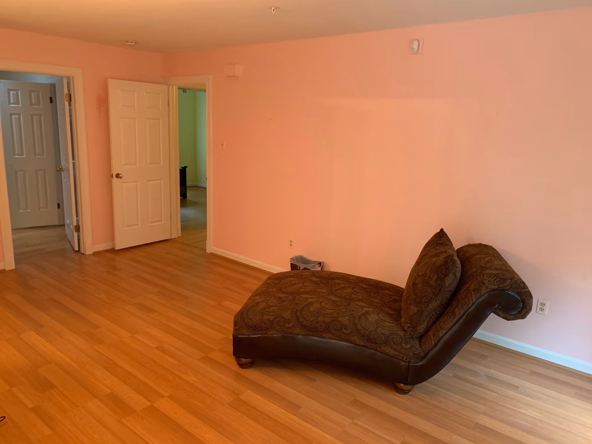 Sparsely furnished room with pink walls and hardwood floors featuring a brown chaise lounge and open doorways to adjacent rooms.